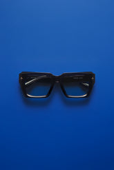 Black square eyeglass frames with dark lenses, photographed flat against a solid blue background.