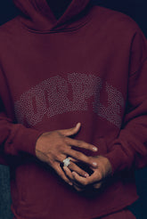 Person wearing a maroon hoodie with a logo, hands clasped together.
