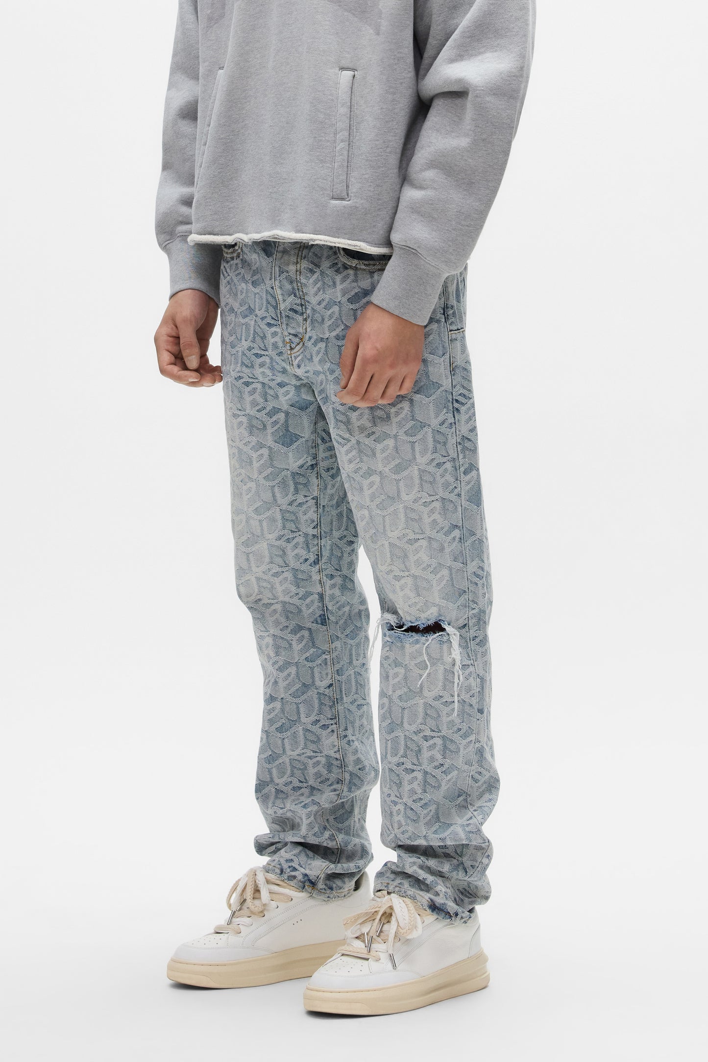 A person wears the PURPLE BRAND P011 Jacquard Monogram light grey sweatshirt, mid-rise ripped knee straight leg jeans, and white sneakers, standing against a plain white background.