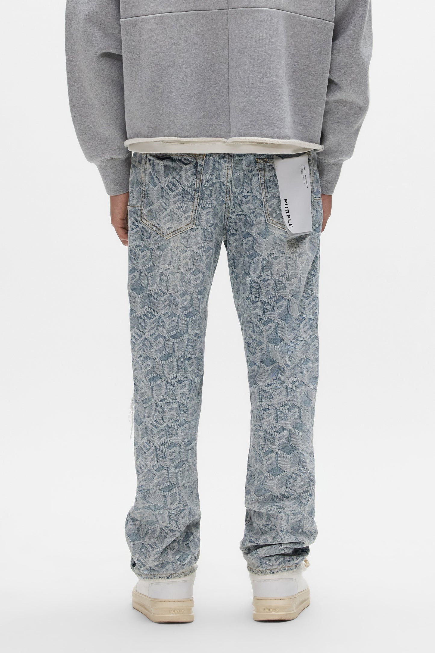 A person faces away wearing PURPLE BRAND P011 Jacquard Monogram light blue mid rise jeans with a white back pocket tag, paired with a grey sweatshirt and white sneakers, set against a plain white background.