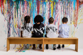 Four children sitting on a bench in front of a colorful wall with splashes of paint.